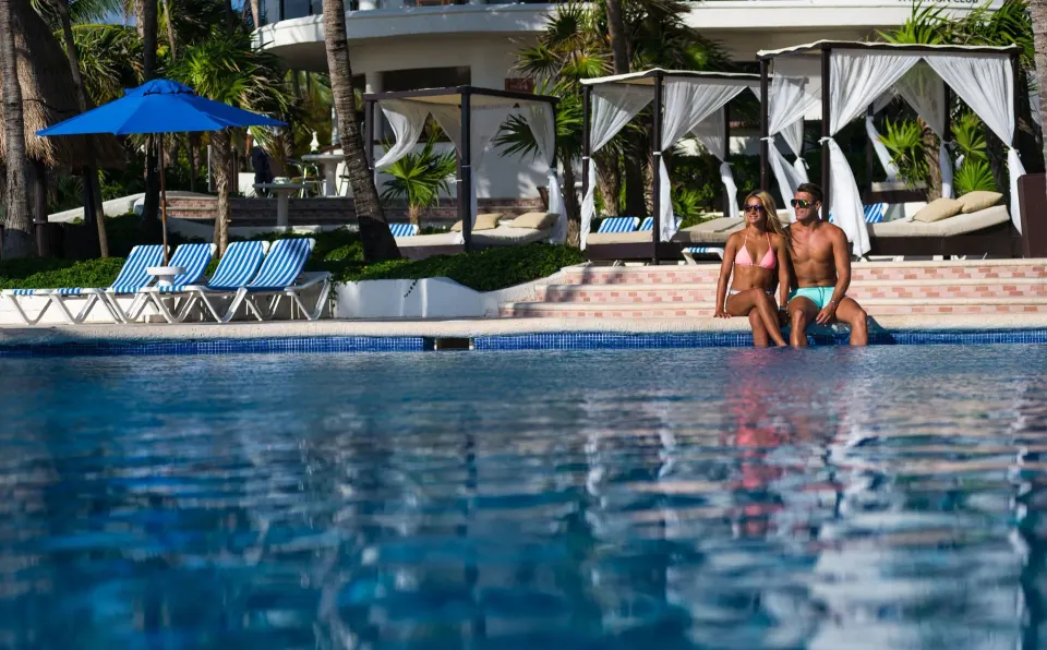 Couple sitting at the pool edge with cabanas behind at The Reef Playacar Beach Resort & Spa