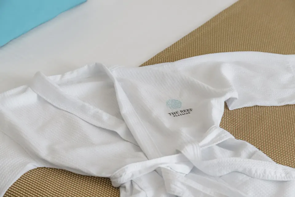 White hotel robe embroidered with The Reef logo on a bed at The Reef Playacar Beach Resort & Spa