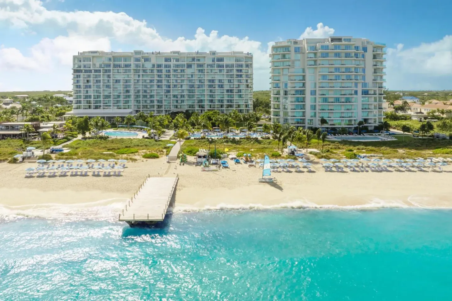Aerial beachfront view of The Ritz-Carlton, Turks & Caicos with towers, beach chairs, pier, and turquoise water.