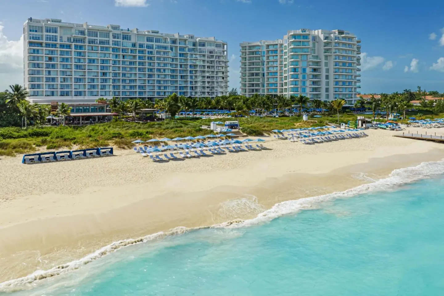 Beachfront view of The Ritz-Carlton, Turks & Caicos with resort towers, lounge chairs, sand, and turquoise surf.