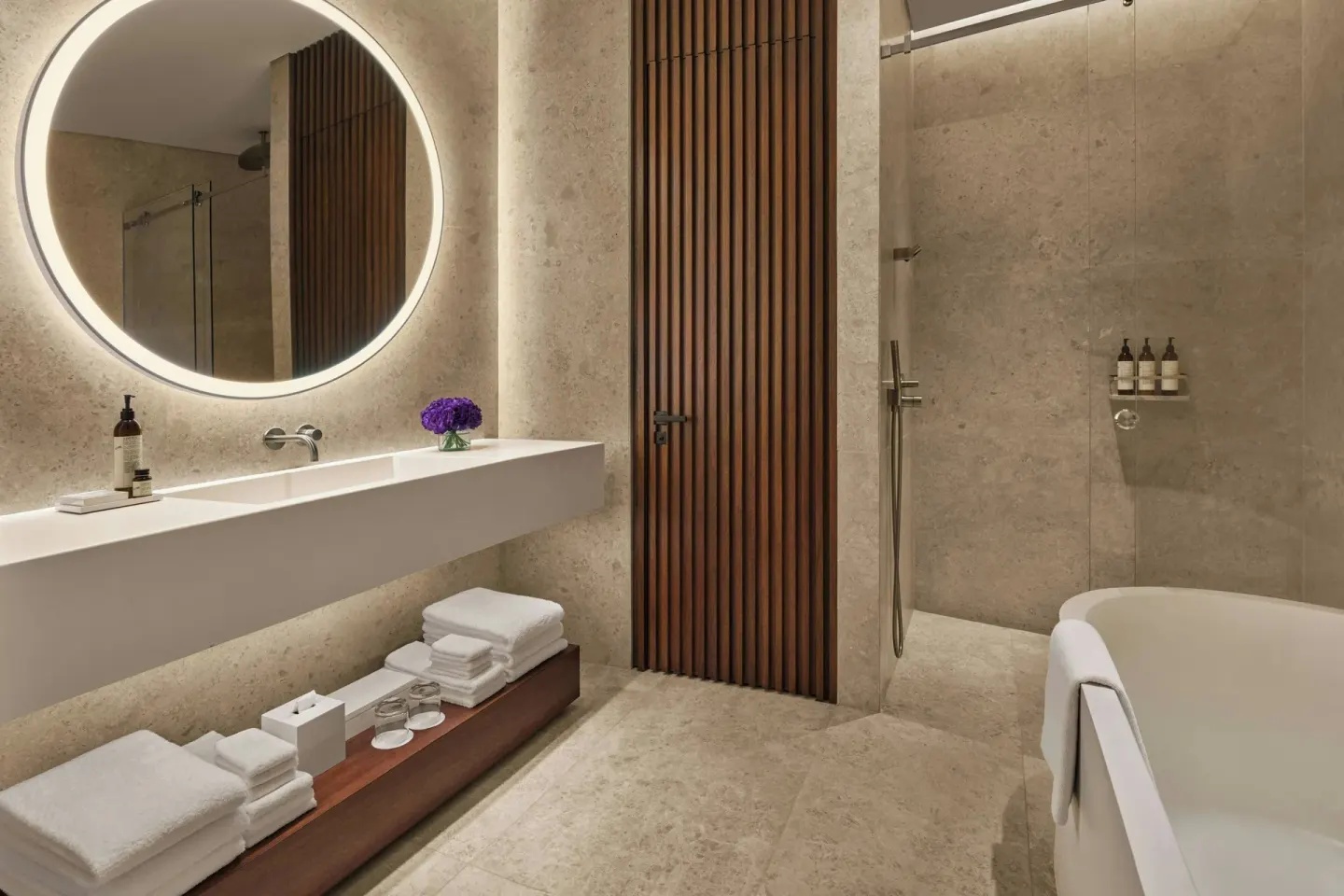 Stone bathroom with round lit mirror and tub at The Riviera Maya EDITION At Kanai.
