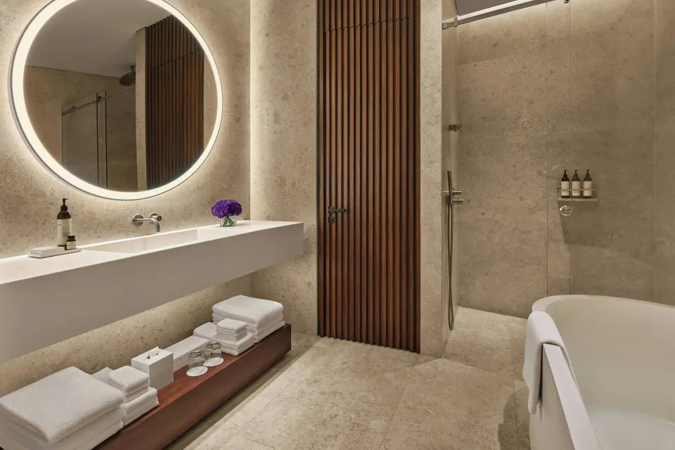 Stone bathroom with round lit mirror and tub at The Riviera Maya EDITION At Kanai.
