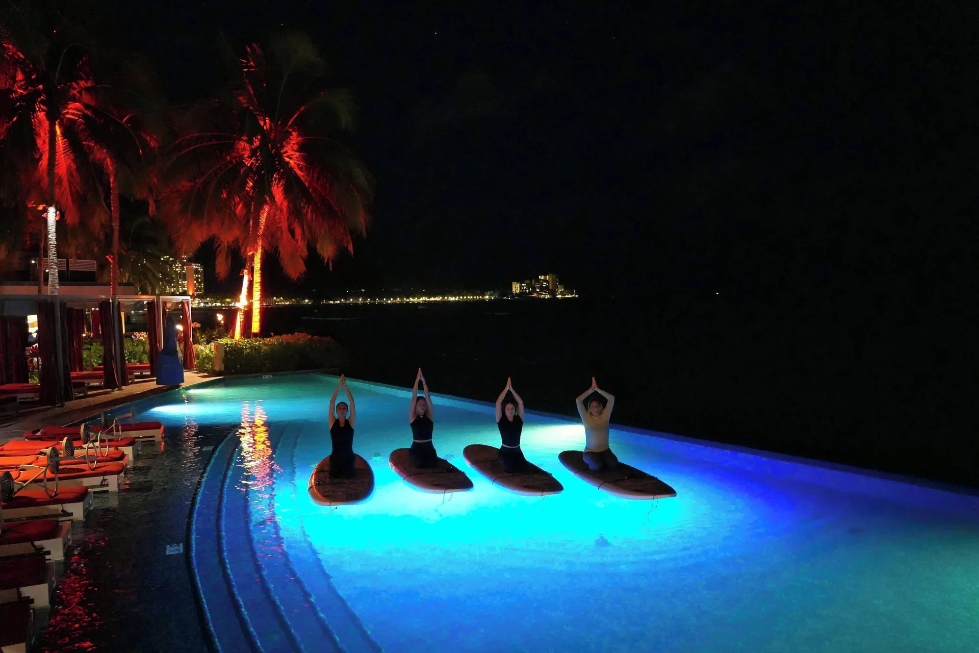 Guests do paddleboard yoga in the illuminated pool at night at The Royal Hawaiian, Waikiki.
