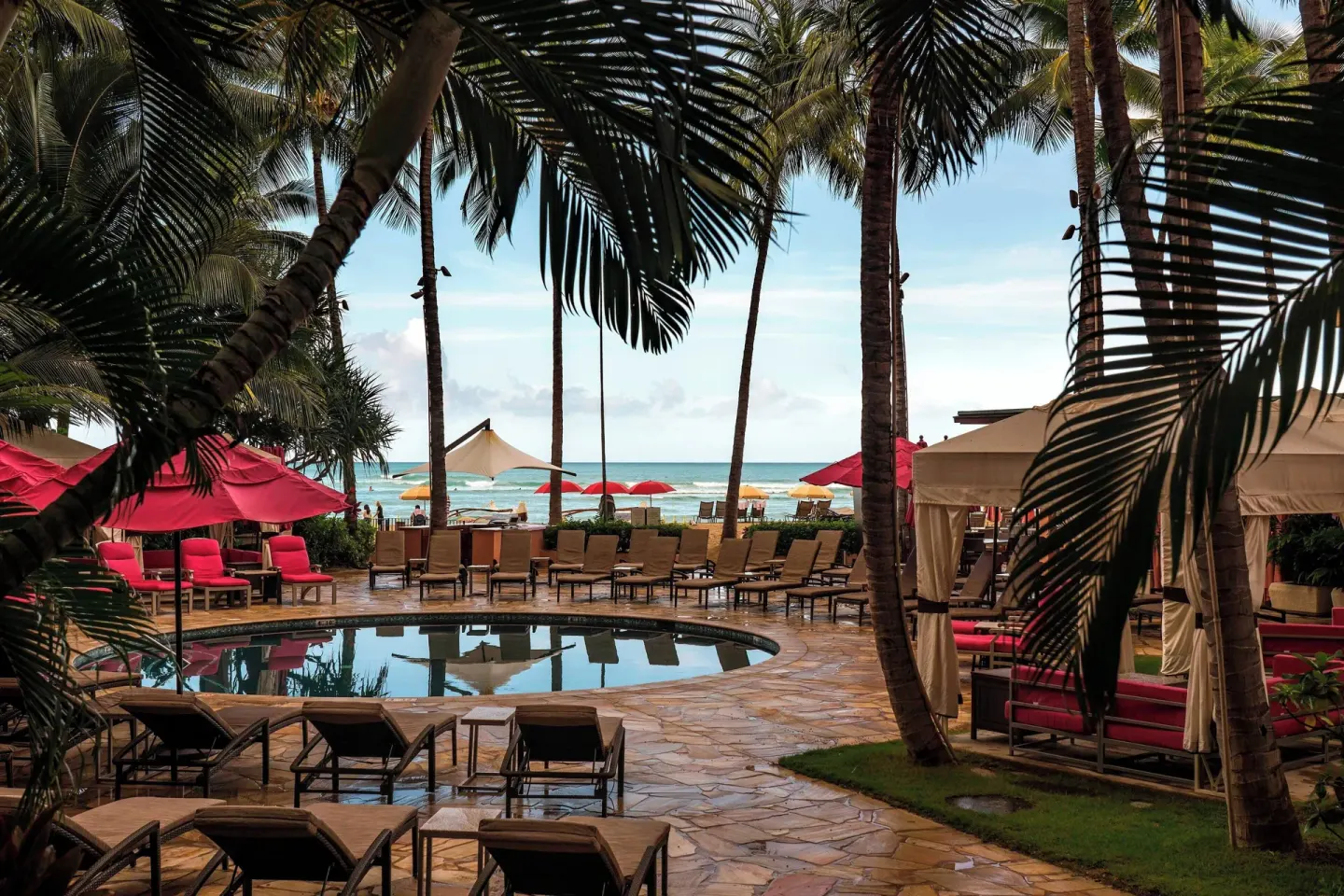 Palm-shaded pool deck with loungers and ocean beyond at The Royal Hawaiian, Waikiki.