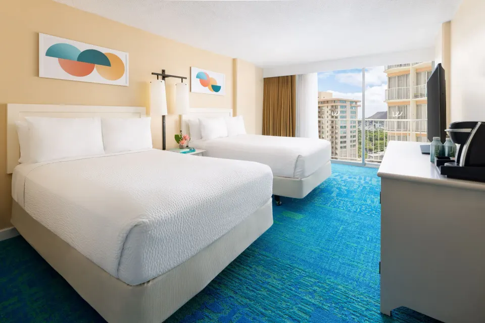 Two white beds and a window with city buildings in a City View room at The Twin Fin Hotel.