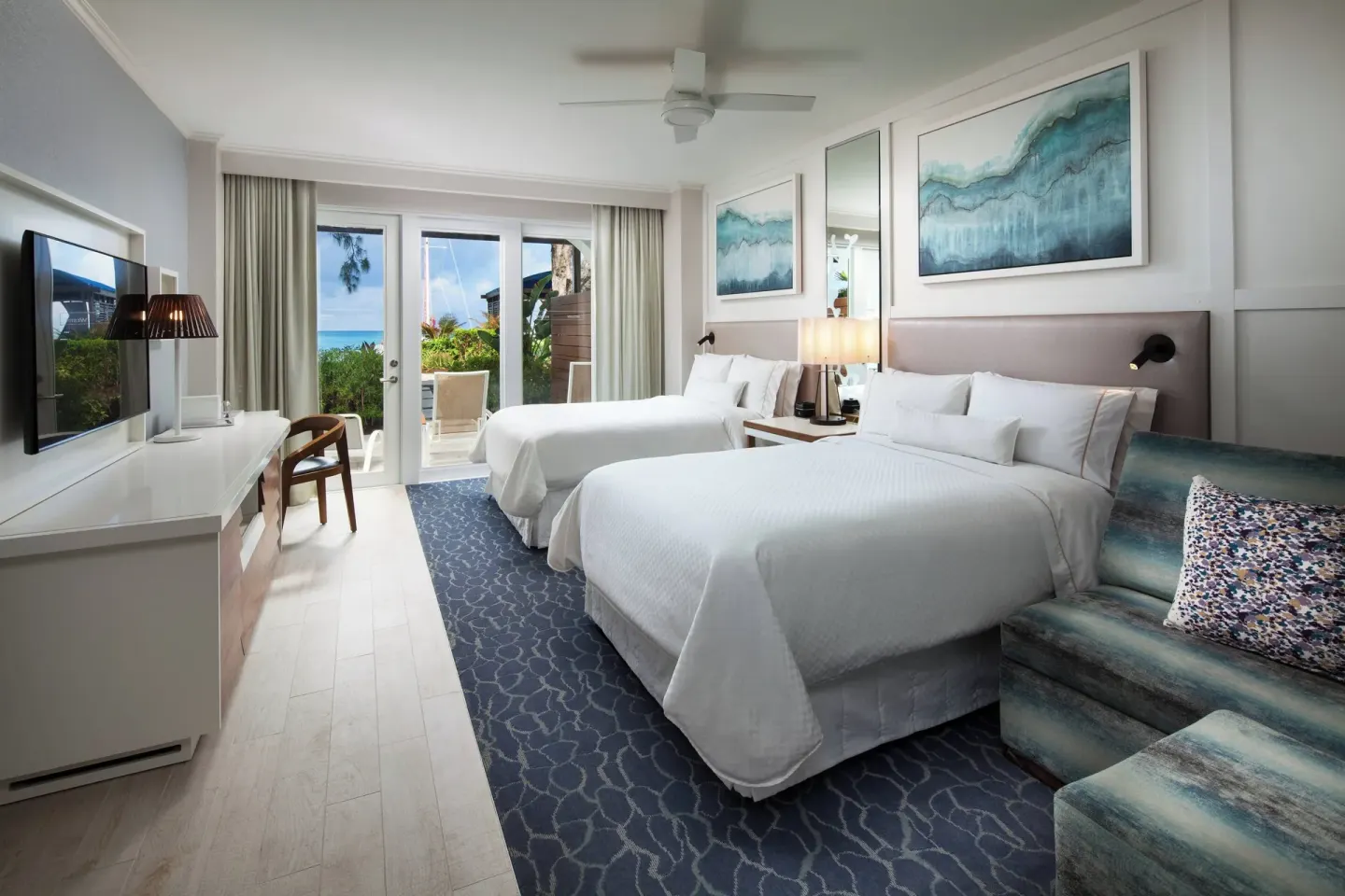Two queen beds and a sofa by patio doors in a room at The Westin Grand Cayman Seven Mile Beach Resort & Spa.