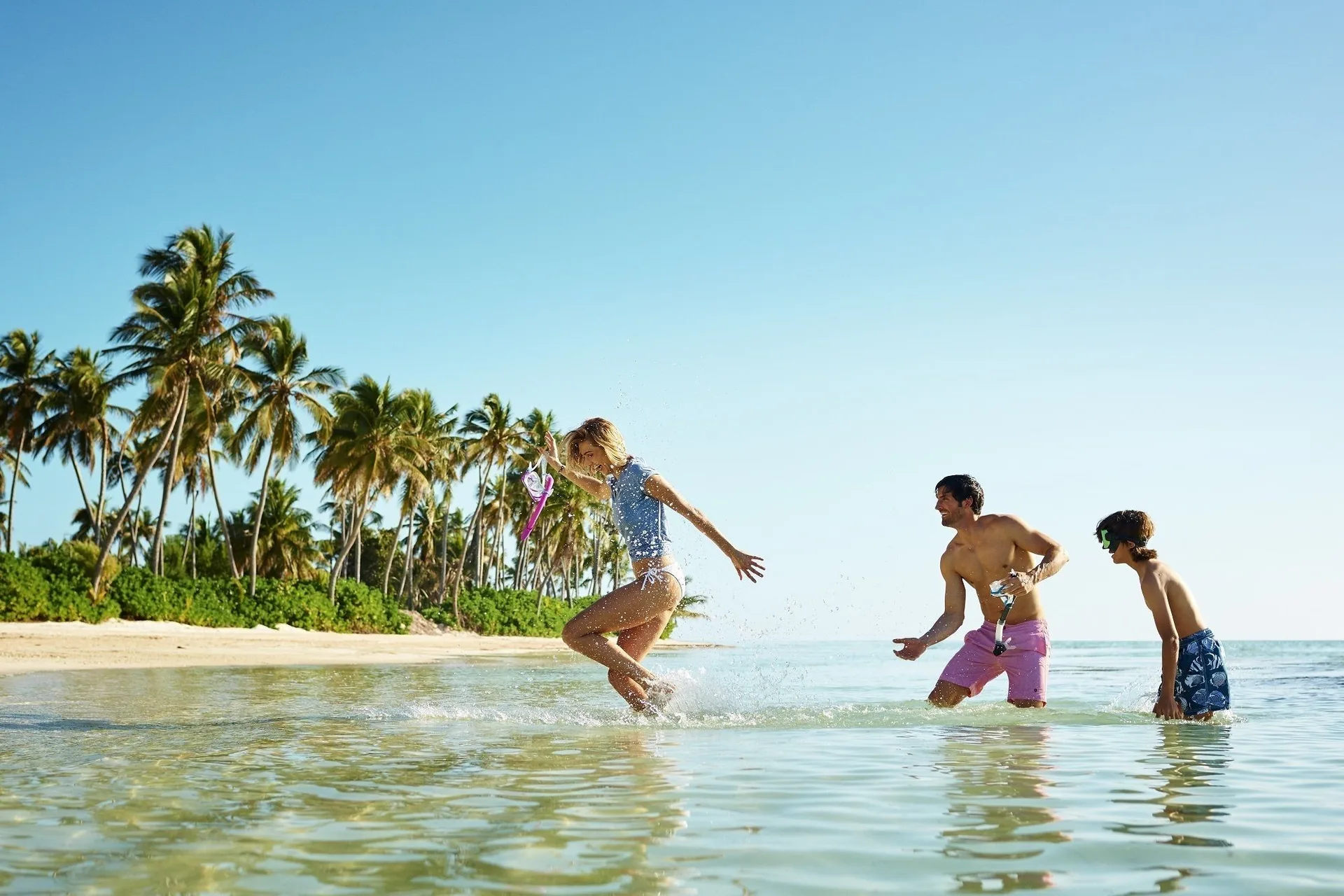 Family playing in shallow ocean water beside a palm-lined beach at Tortuga Bay Hotel.