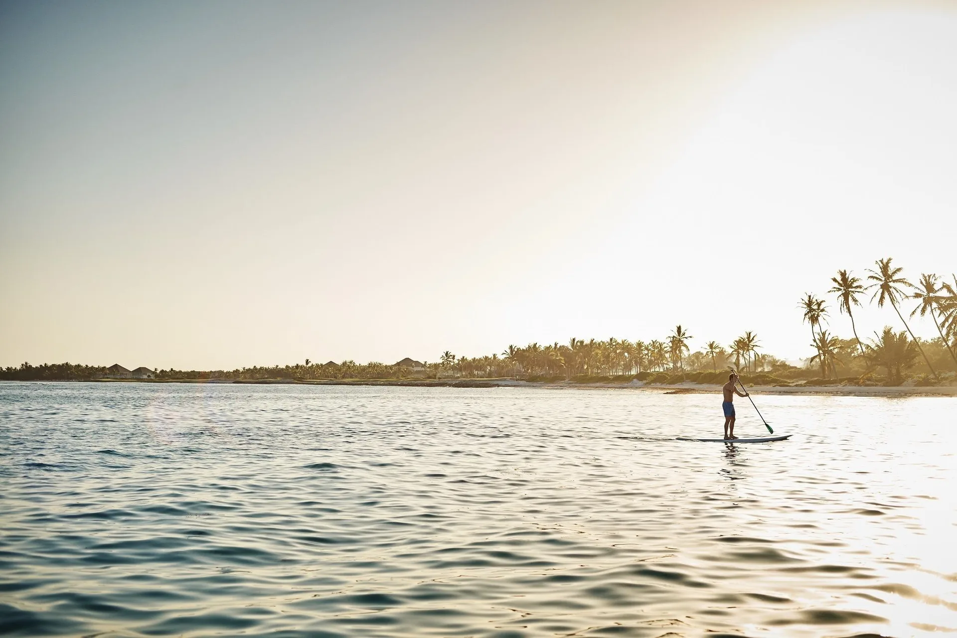 Paddleboarder on calm coastal water near Tortuga Bay Hotel at sunset.