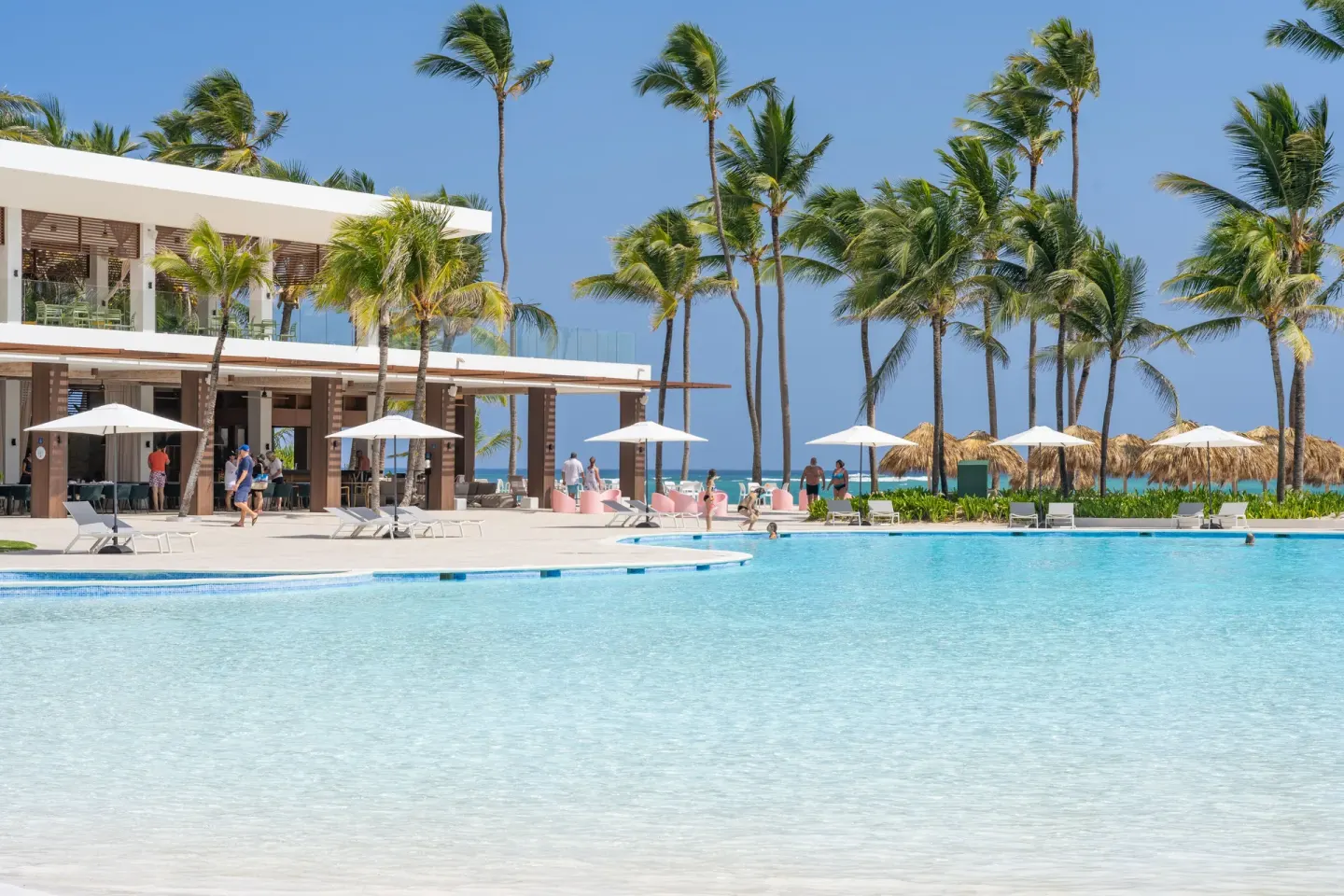 Sunny pool beside palm trees, umbrellas and a beachfront building at Tropical Deluxe Princess.