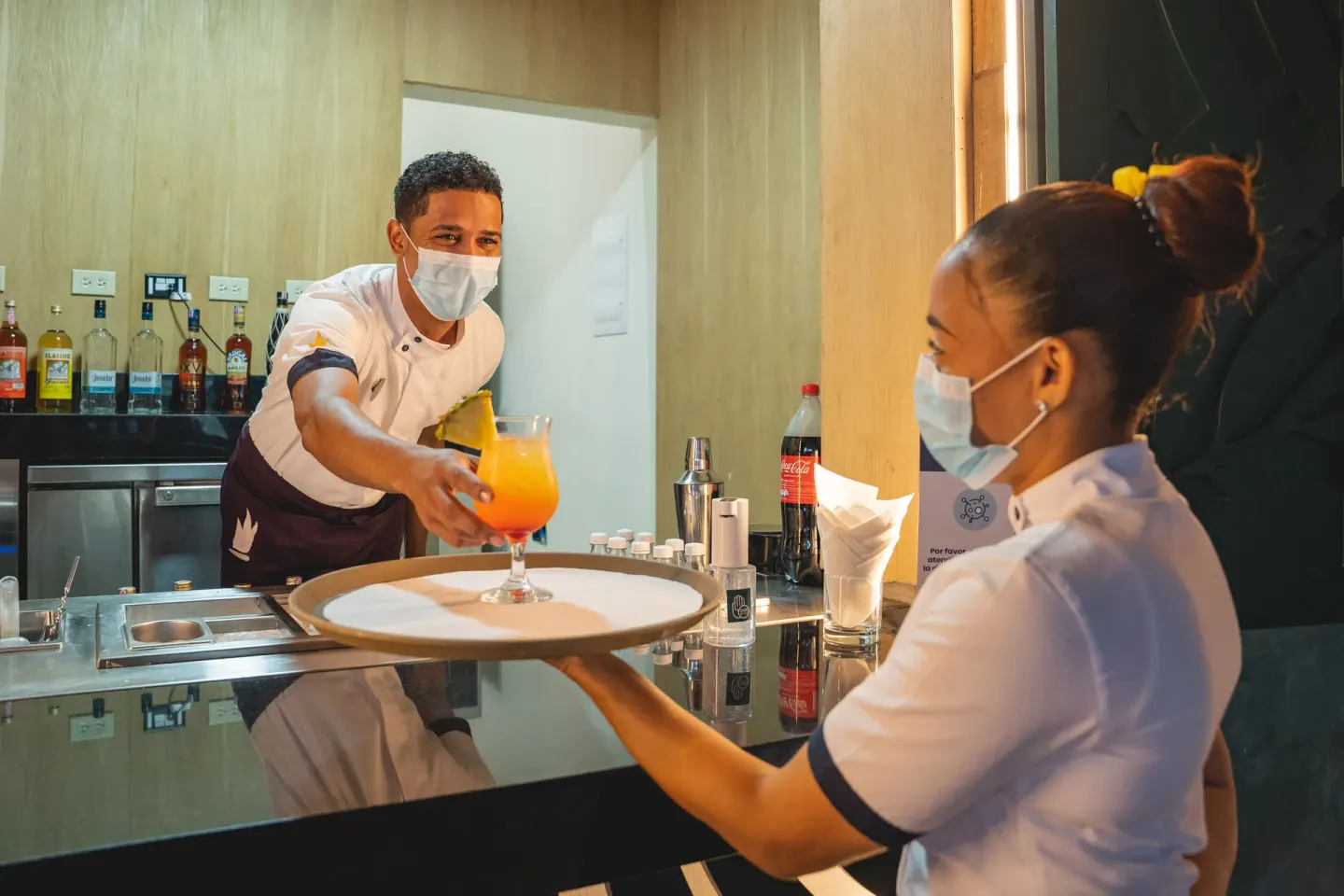 Masked bartender handing a tropical drink to a staff member at Tropical Deluxe Princess.