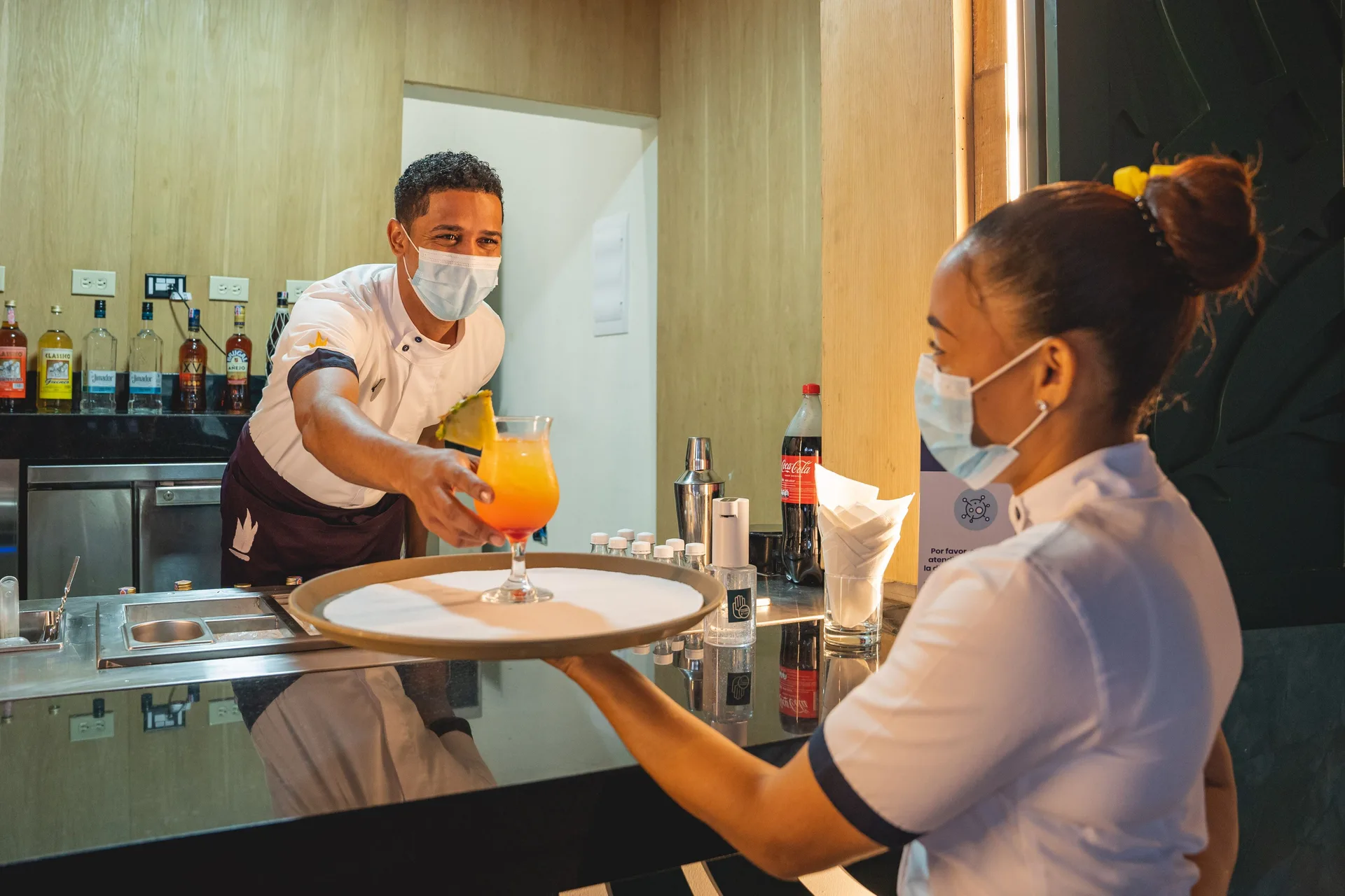 Masked bartender handing a tropical drink to a staff member at Tropical Deluxe Princess.