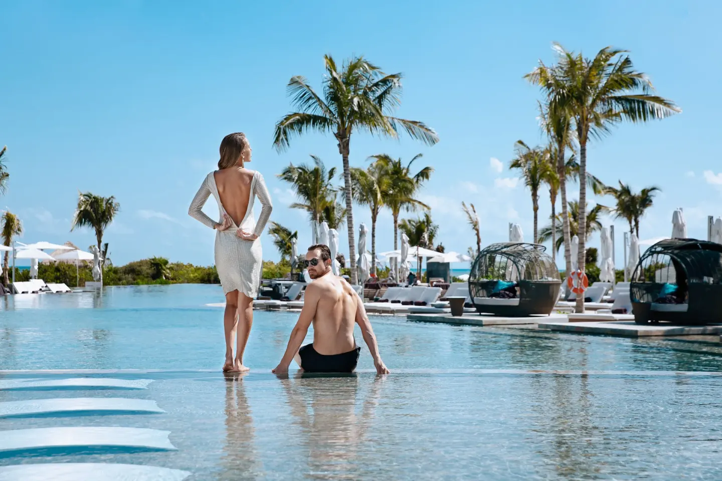 Couple standing and sitting in the pool at TRS Coral Hotel with palms, loungers, and cabanas behind them.
