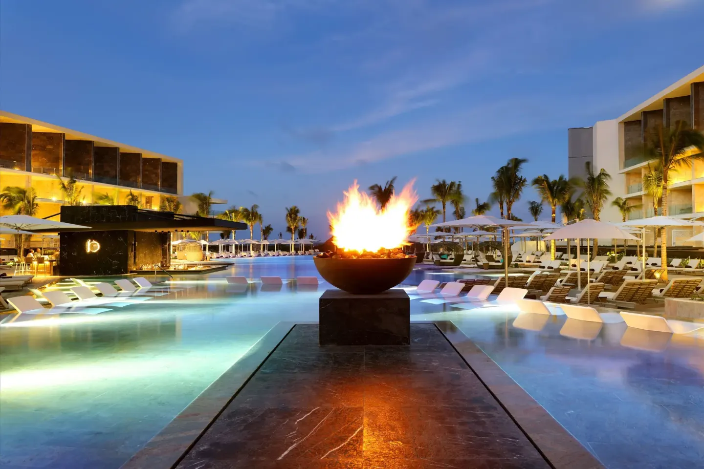 Evening pool scene at TRS Coral Hotel with a fire bowl, lit water, loungers, and umbrellas.