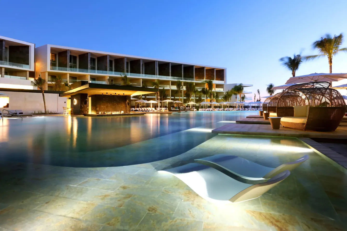 Twilight pool with submerged lounger, woven cabanas and lit bar at TRS Coral Hotel.
