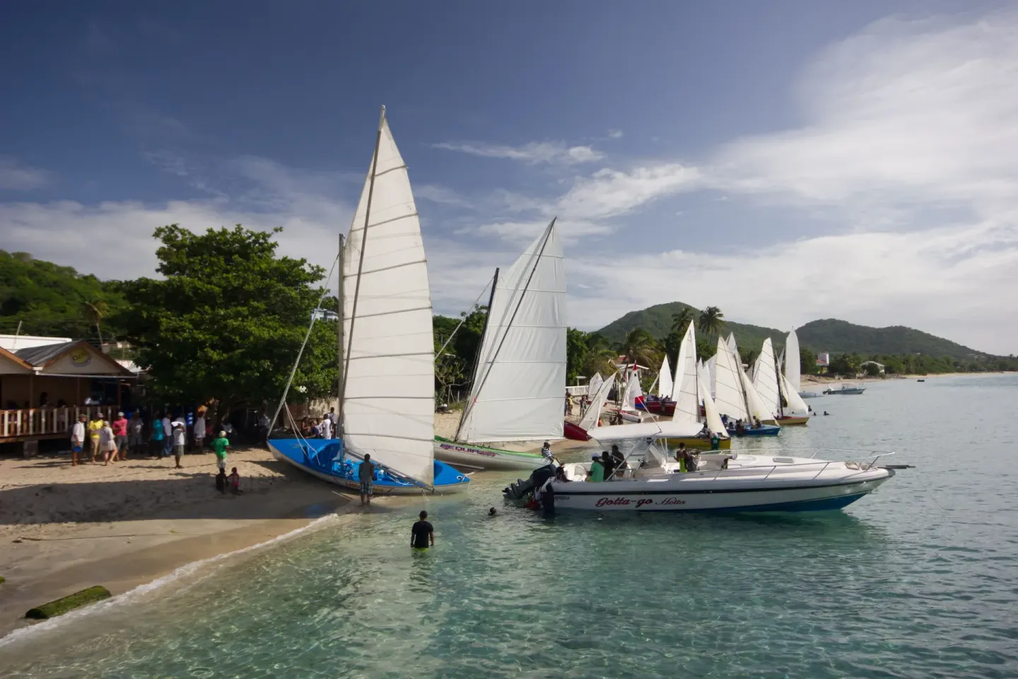 Sailboats line a sandy beach with people by the water near True Blue Bay Resort.