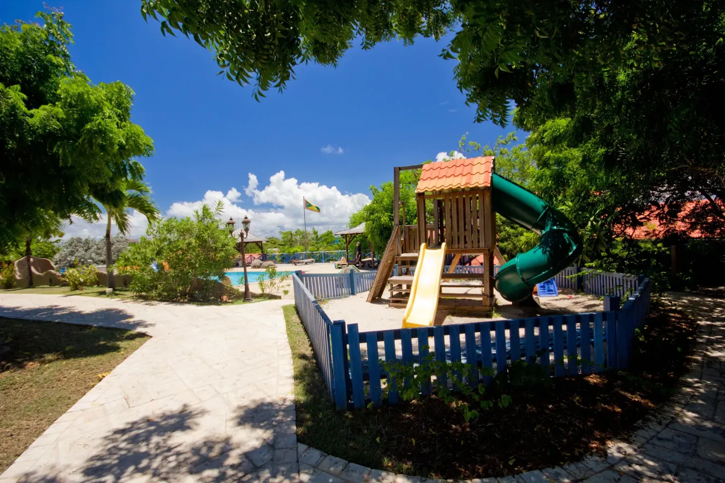 Fenced playground with slides beside a pool and garden paths at True Blue Bay Resort.