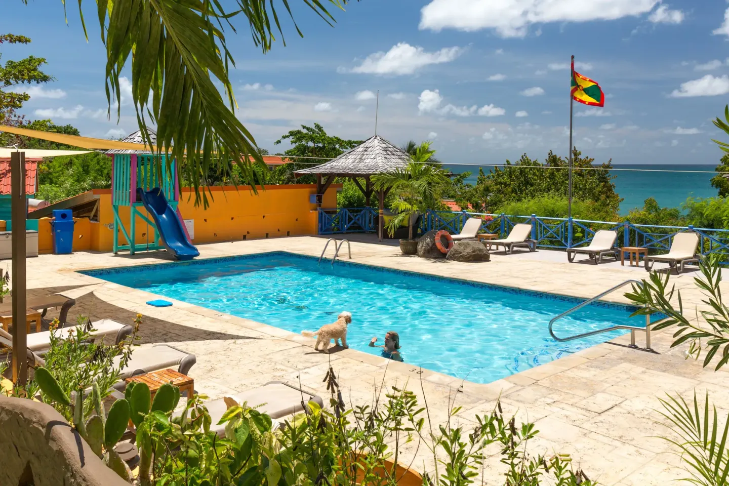 Pool with loungers, slide, sea view, and Grenada flag at True Blue Bay Resort.