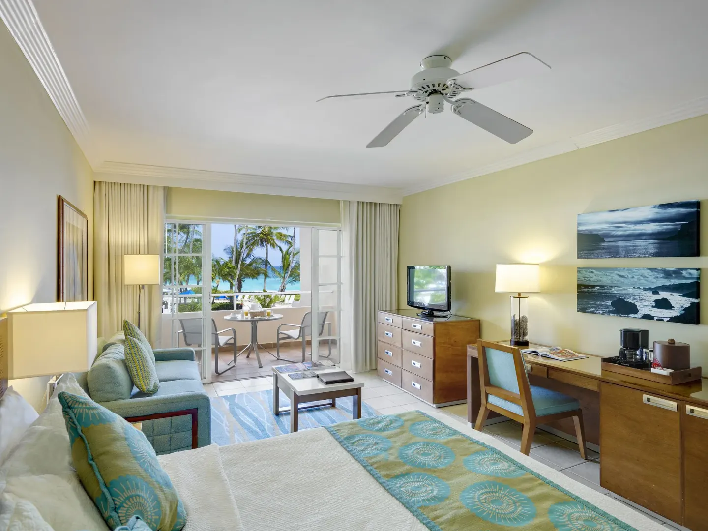 Junior suite at Turtle Beach with bed, sofa, desk, balcony, and ocean beyond palm trees.