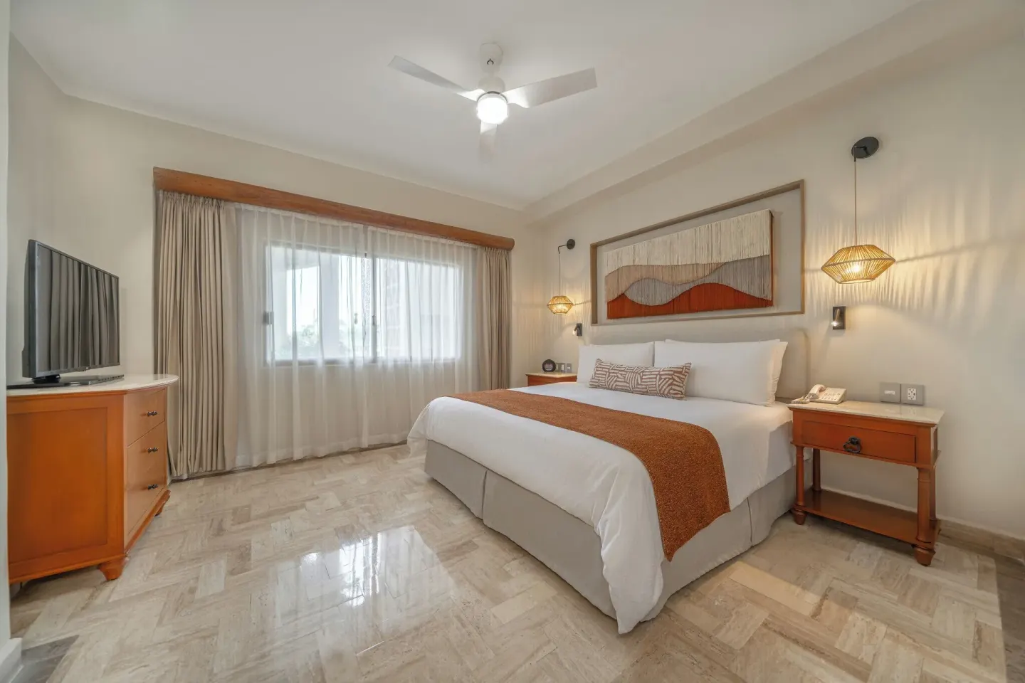 King bed guest room with TV, marble floor, and curtained window at Velas Vallarta Suites Resort.