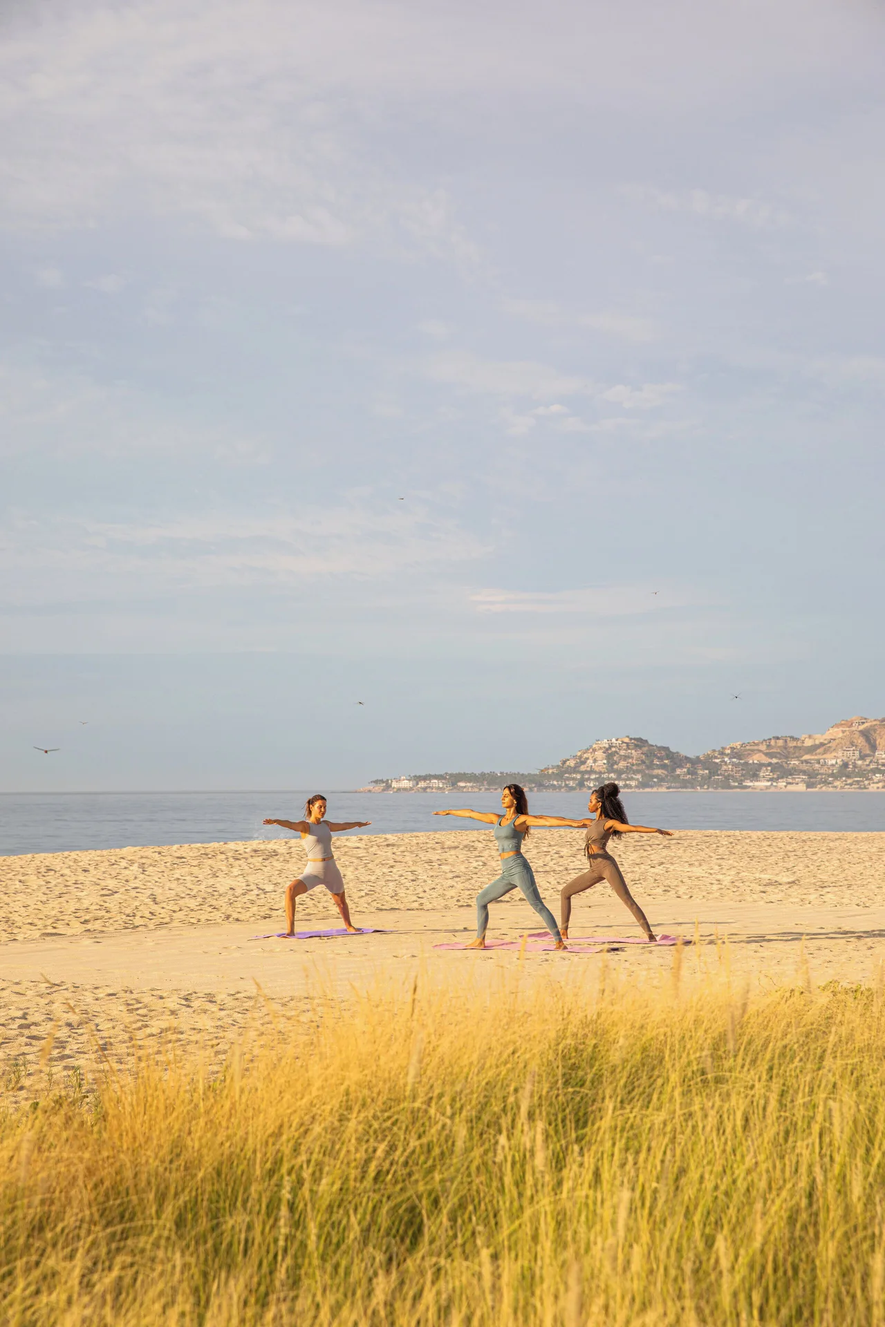 Three people practice yoga on the beach with the sea and coastline beyond Viceroy Los Cabos.