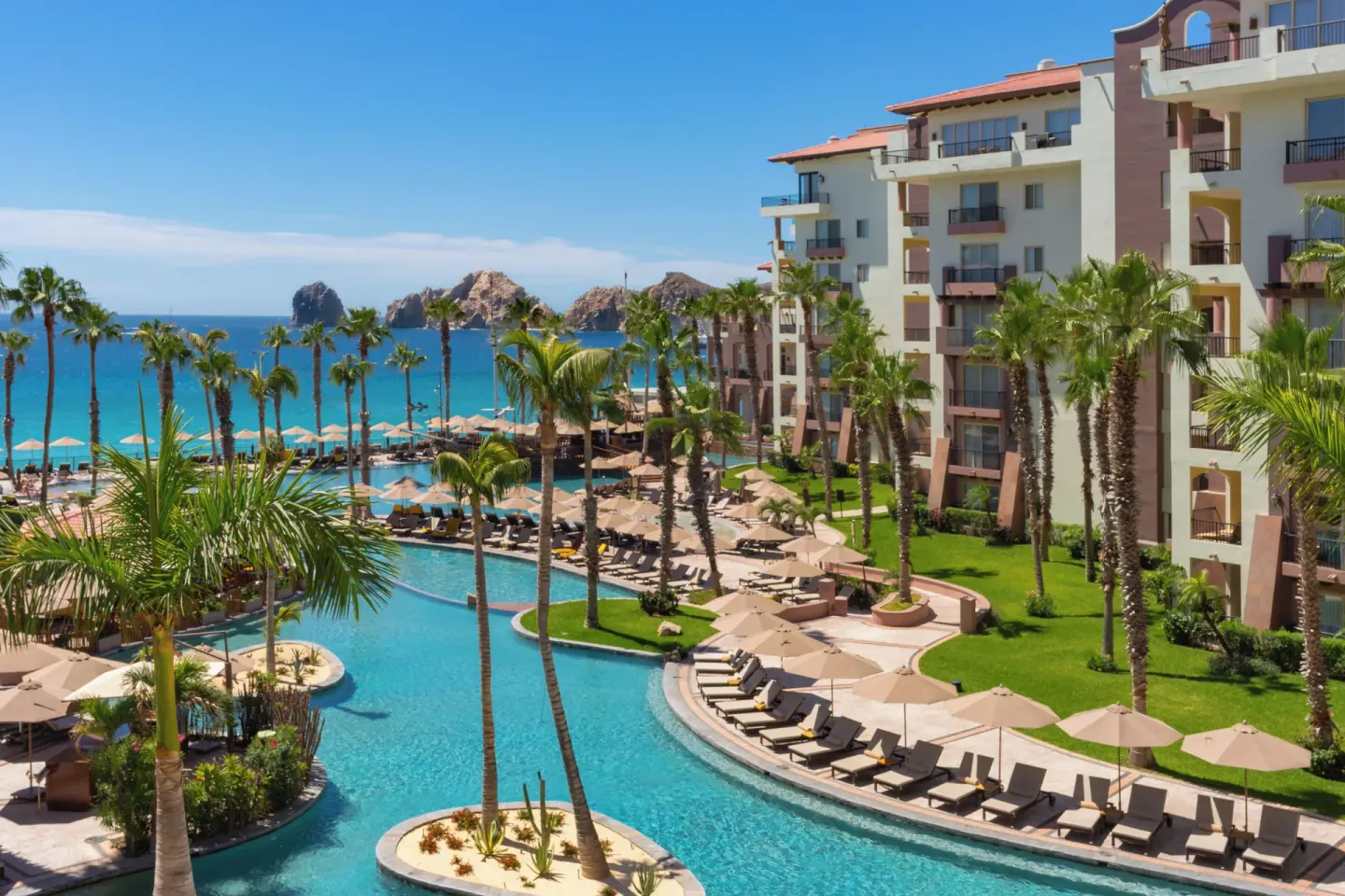 Curving pool, loungers, palms, and ocean at Villa del Arco Beach Resort & Spa Cabo San Lucas.