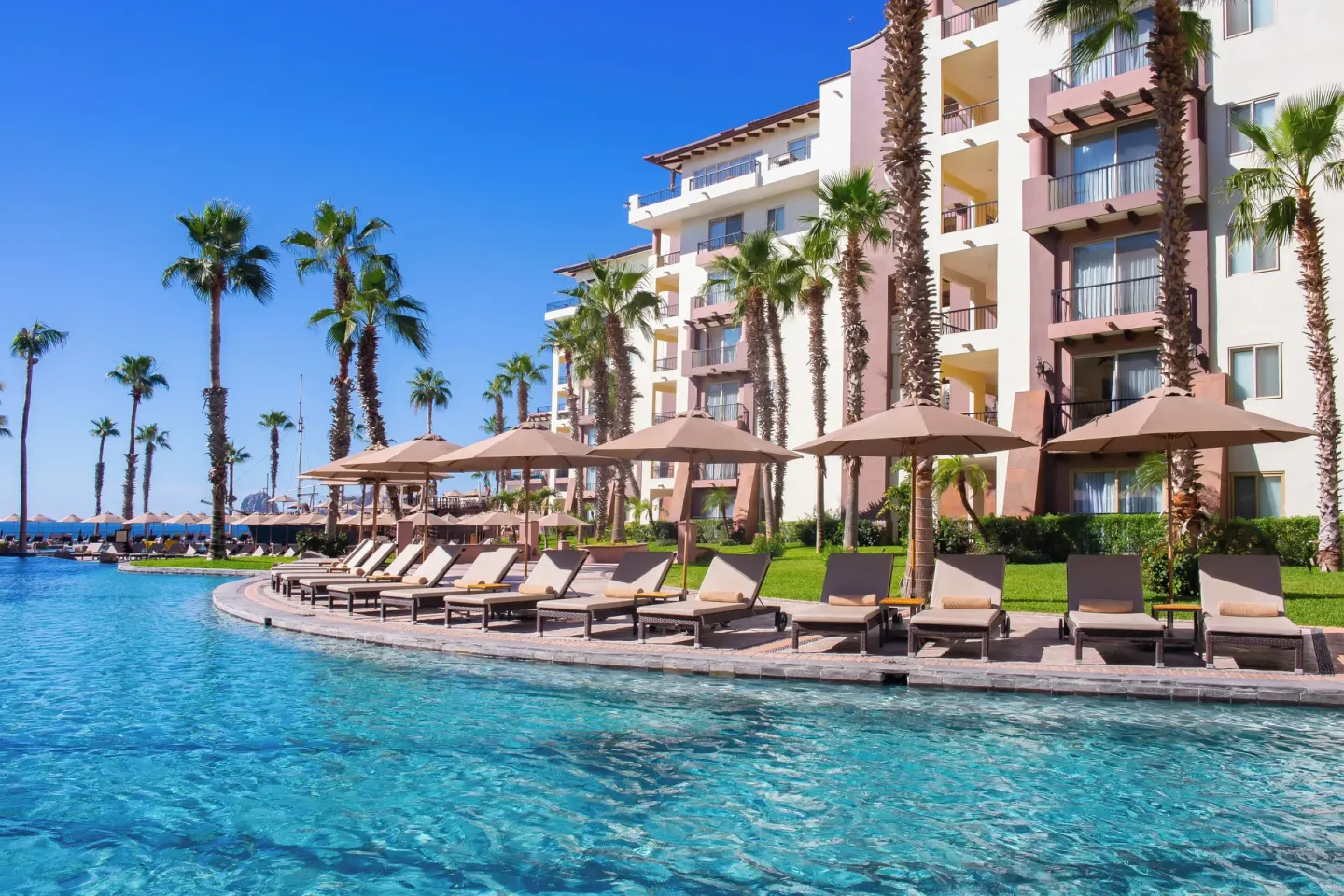 Pool loungers, umbrellas, palms, and resort balconies at Villa del Arco Beach Resort & Spa Cabo San Lucas.
