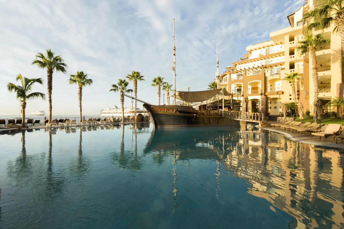 Resort pool at Villa del Arco Beach Resort & Spa Cabo San Lucas with a ship-shaped bar, palms, and ocean beyond.