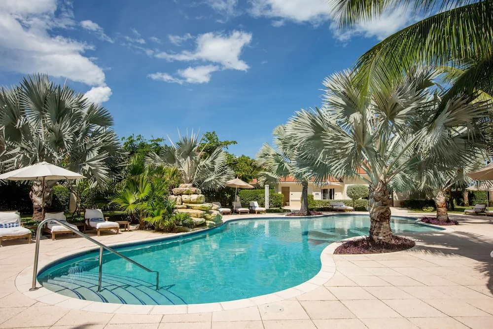A sunny pool with palms, loungers, umbrellas, and steps is shown at Villa del Mar.