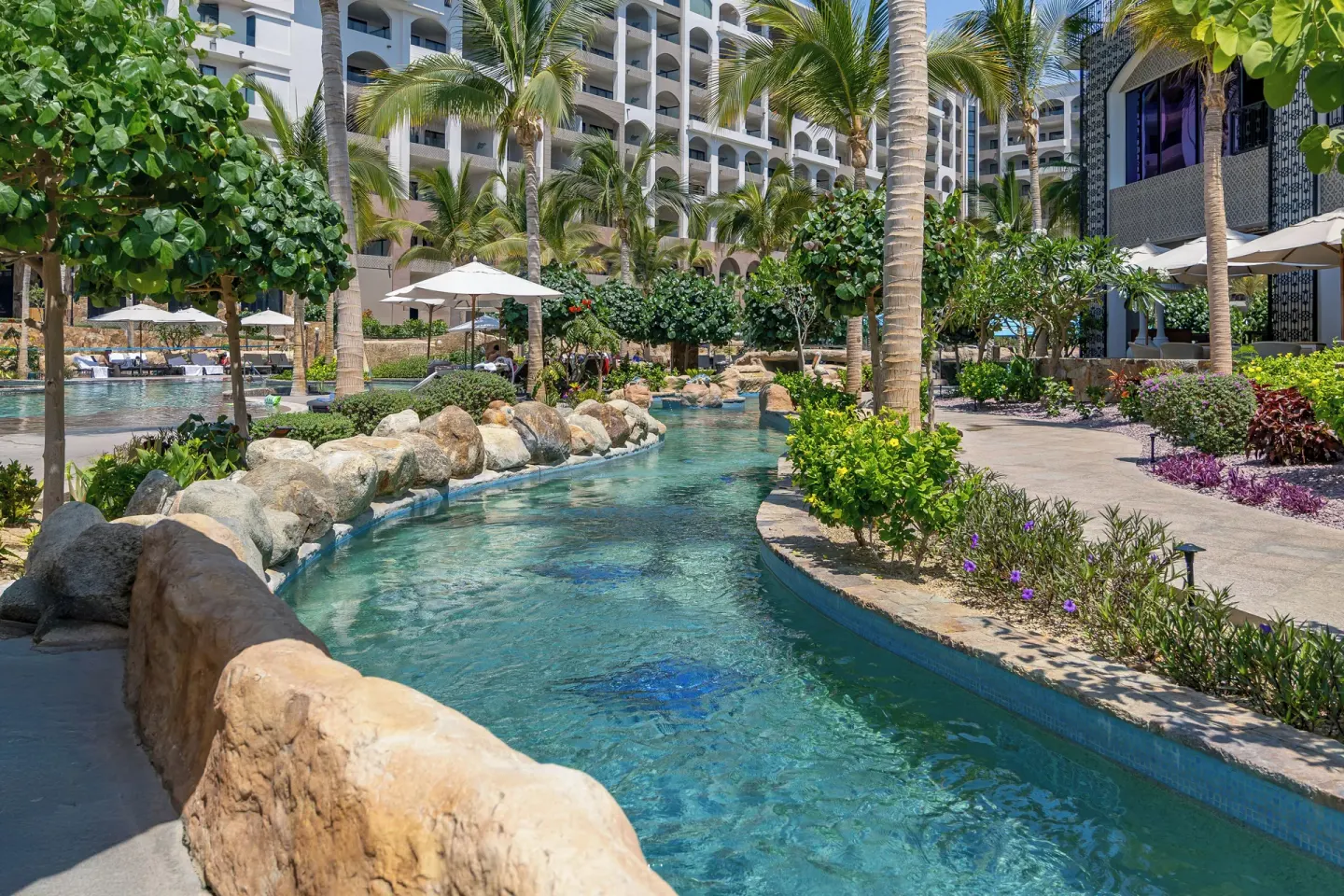 Lazy river garden path with palms and balconies at Villa La Valencia Los Cabos Beach Resort & Spa.