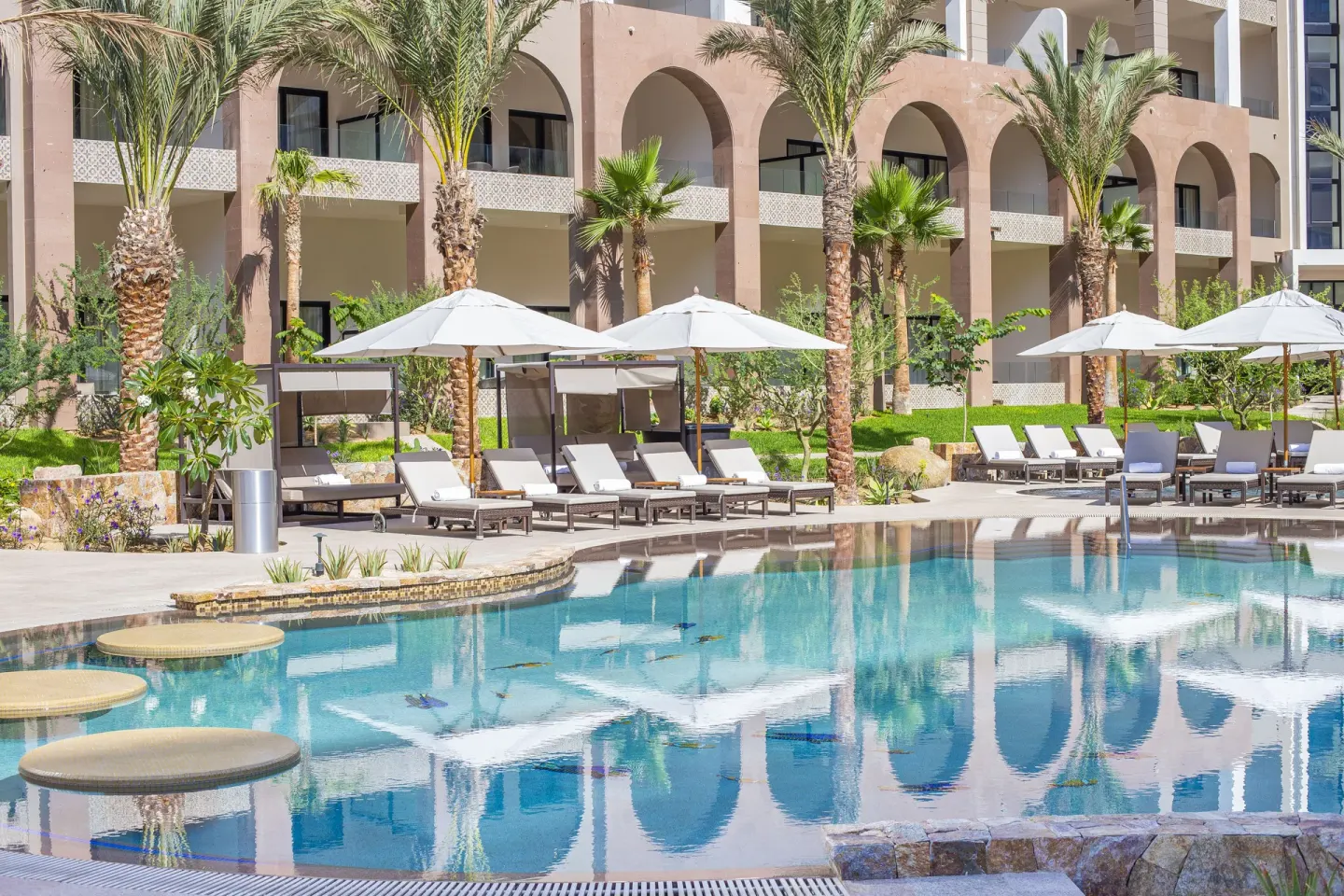 Pool loungers and palms beside arched balconies at Villa La Valencia Los Cabos Beach Resort & Spa.