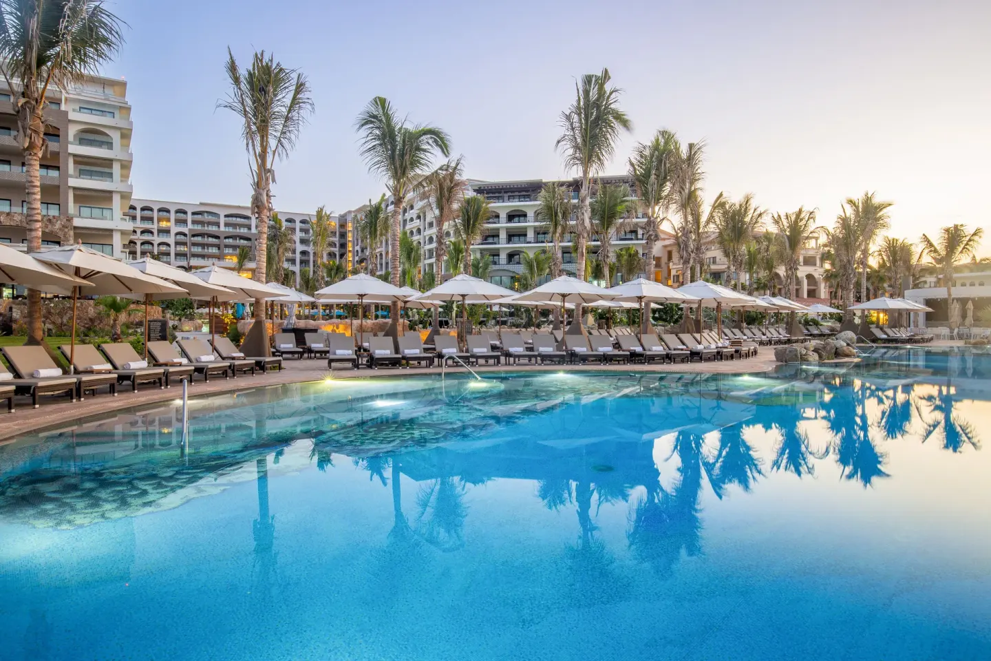 Resort pool at dusk with palms and rows of loungers at Villa La Valencia Los Cabos Beach Resort & Spa.