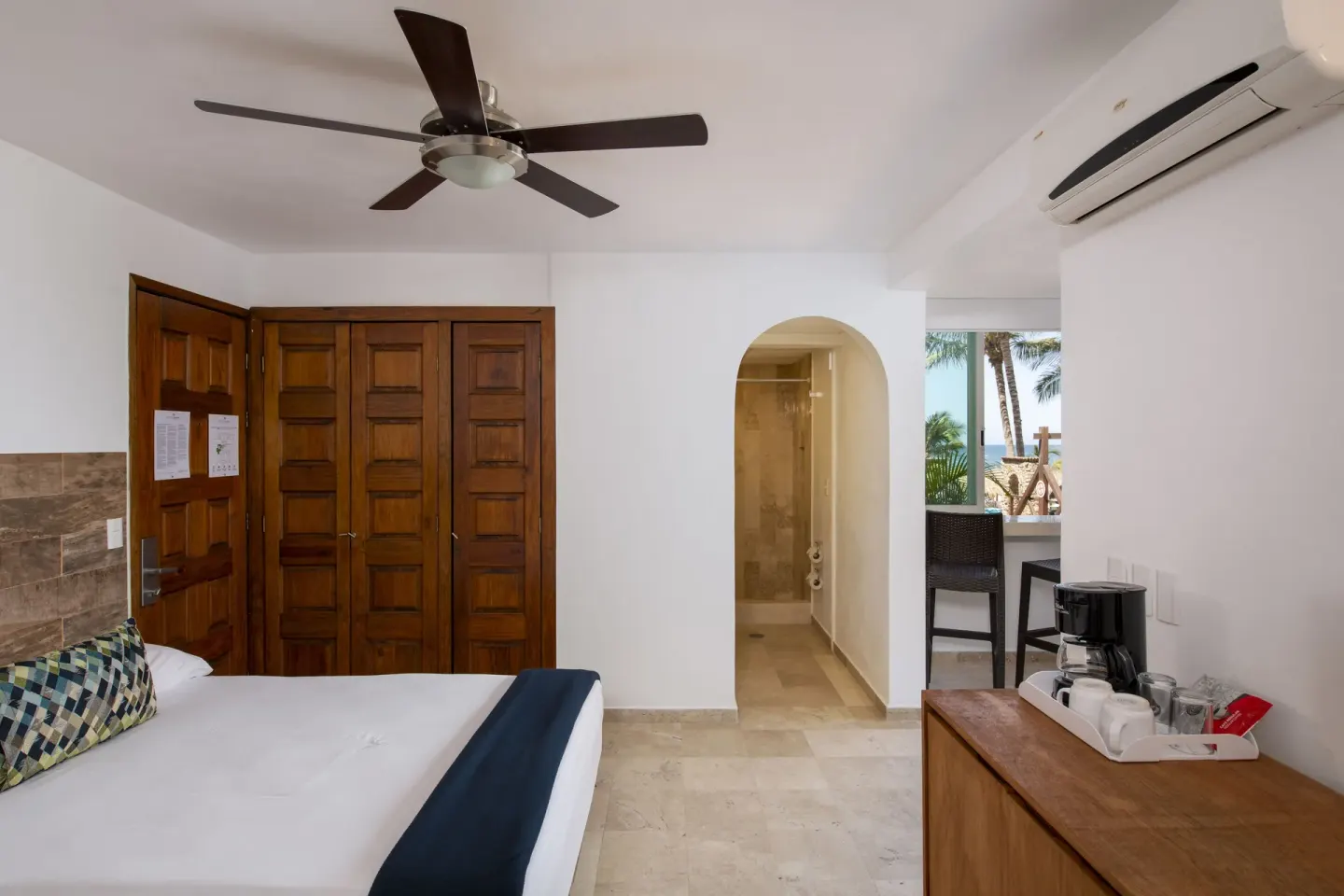 Standard Room at Villa Varadero with a bed, wood doors, coffee maker, and balcony beyond an archway.