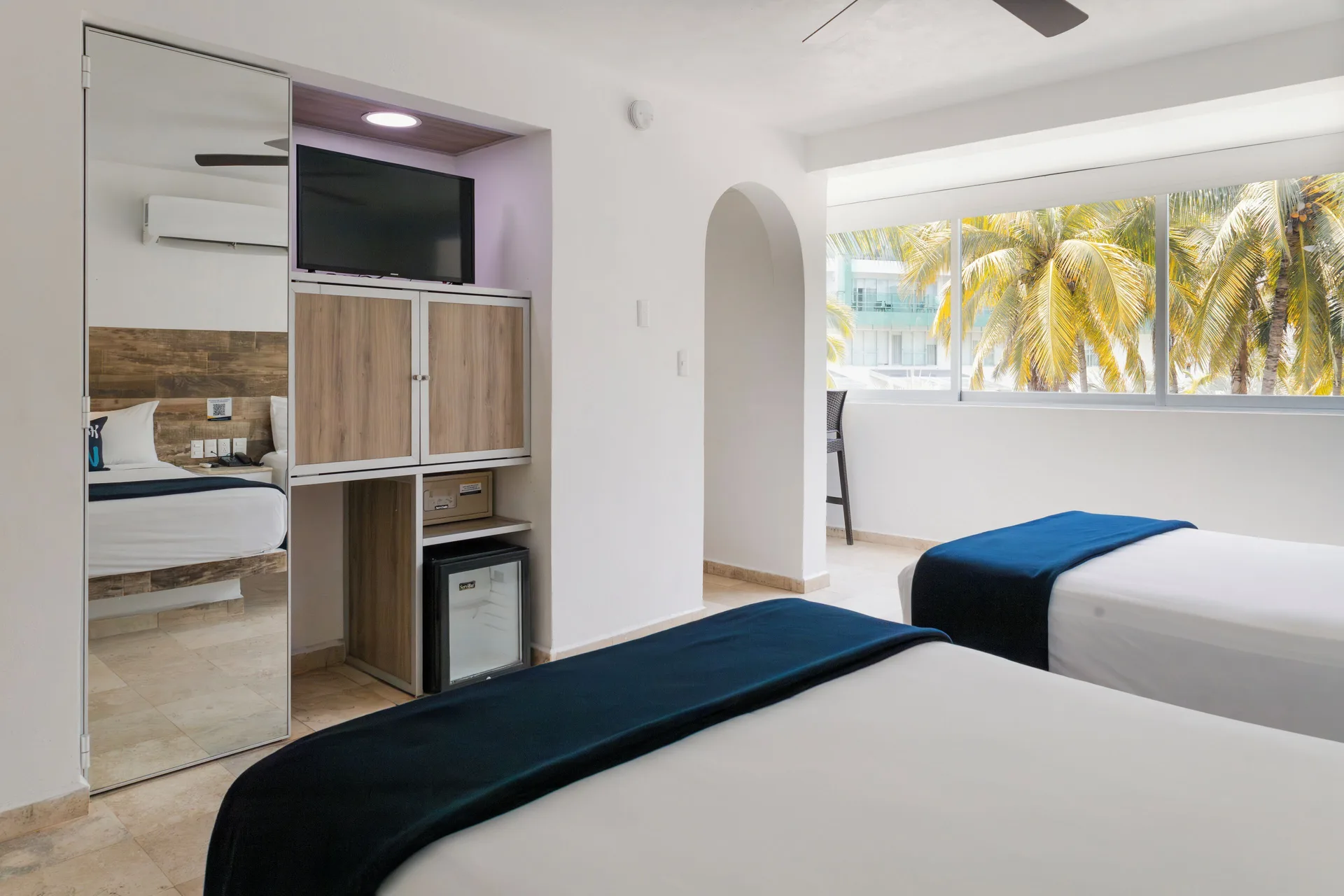 Standard Room at Villa Varadero with two beds, TV cabinet, mirror, mini fridge, and palm-view window.