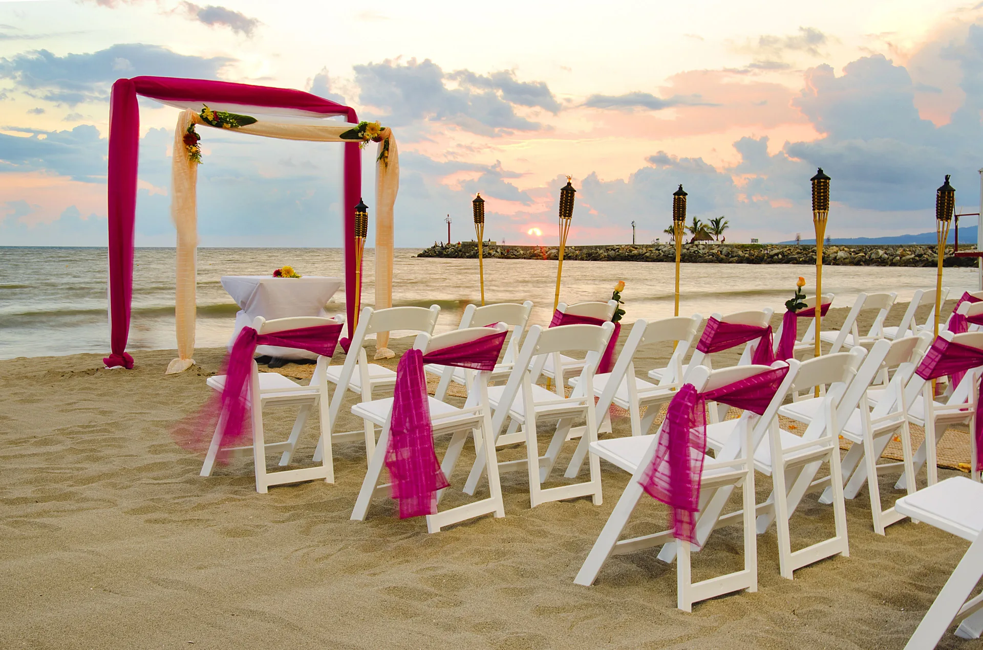 Beach wedding setup with white chairs, pink sashes, torches, and arch at Villa Varadero.