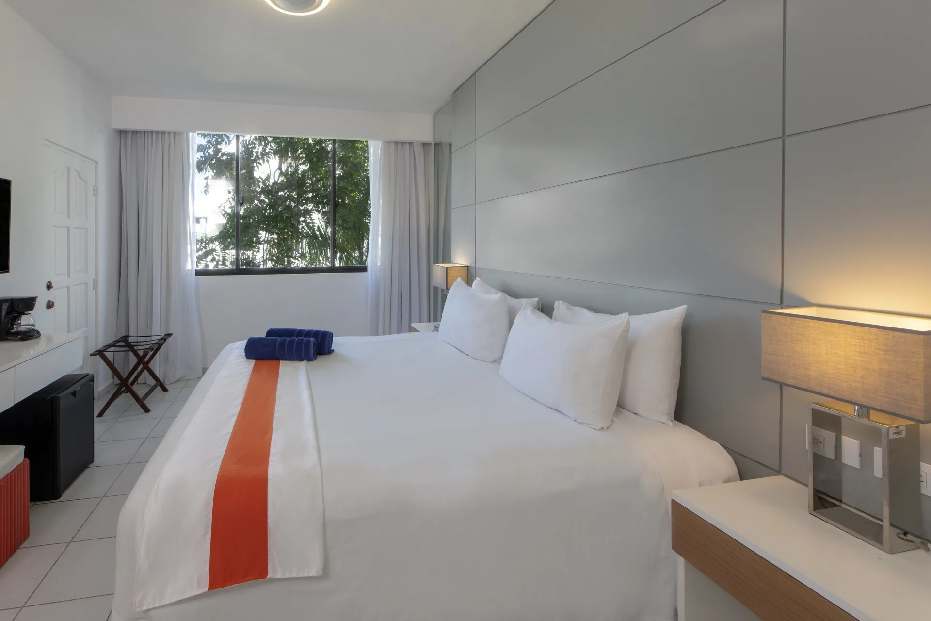 White guest room with a king bed, window, lamps, and tiled floor at Viva Heavens.