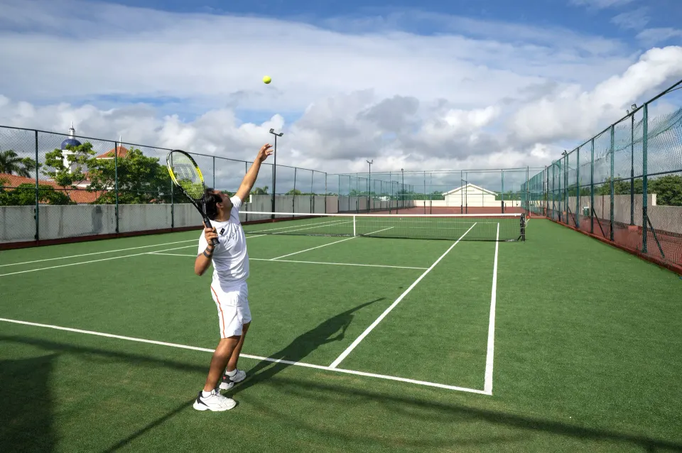 Guest serving a tennis ball on an outdoor court at Viva Maya
