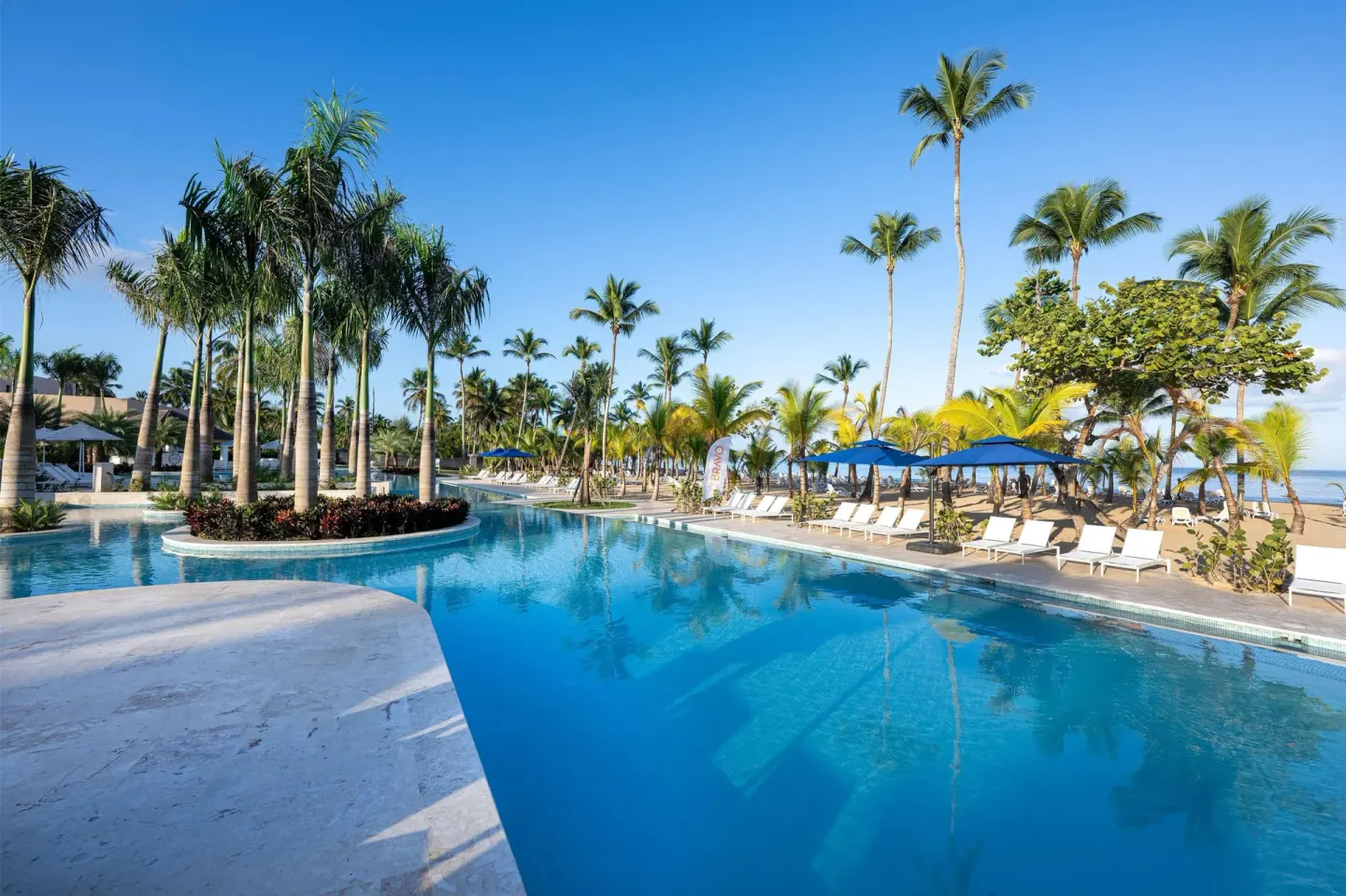 Long pool with palm islands, loungers, blue umbrellas, and beach beyond at Viva Miches.