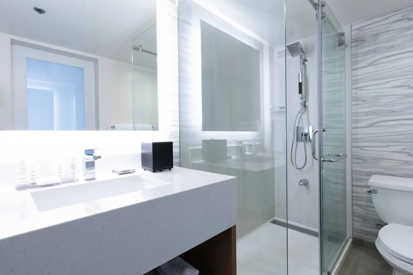 Guest room bathroom at Waikiki Beach Marriott Resort & Spa with vanity, lit mirror, glass shower, and toilet.