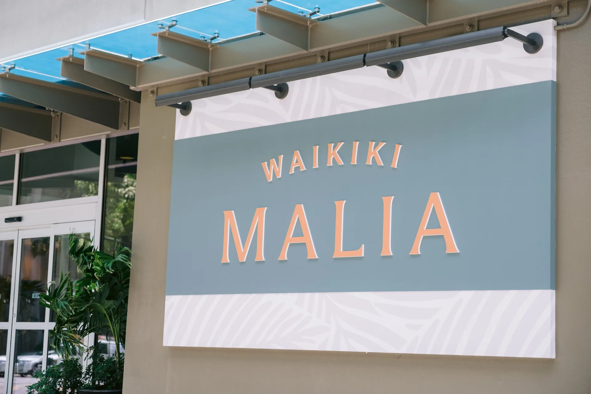 The Waikiki Malia entrance sign is mounted beside glass doors and plants.
