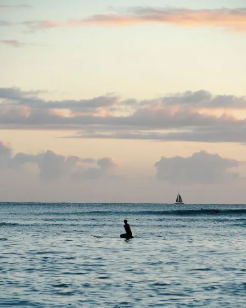 A surfer sits on a board at sunset near Waikiki Malia with a sailboat on the horizon.