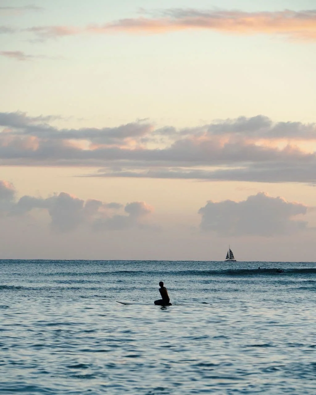 A surfer sits on a board at sunset near Waikiki Malia with a sailboat on the horizon.