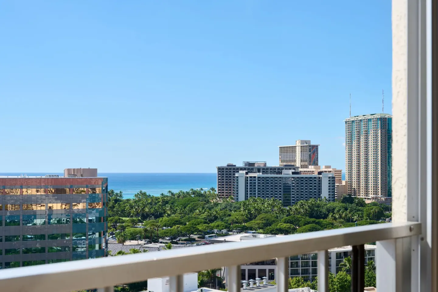 Partial ocean view from a Waikiki Malia balcony with buildings, treetops, and blue water.