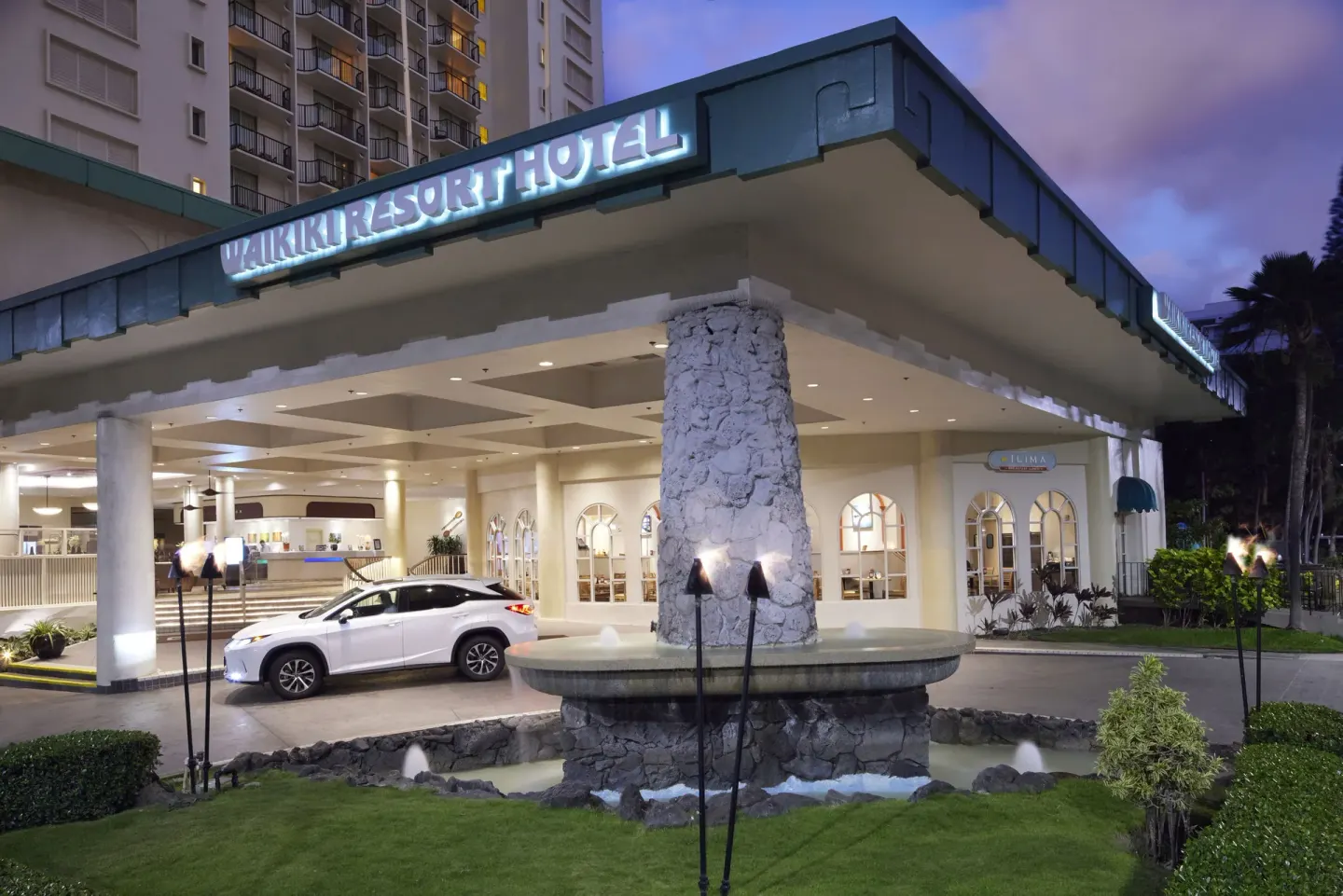 Illuminated entrance canopy and fountain at Waikiki Resort Hotel at dusk.