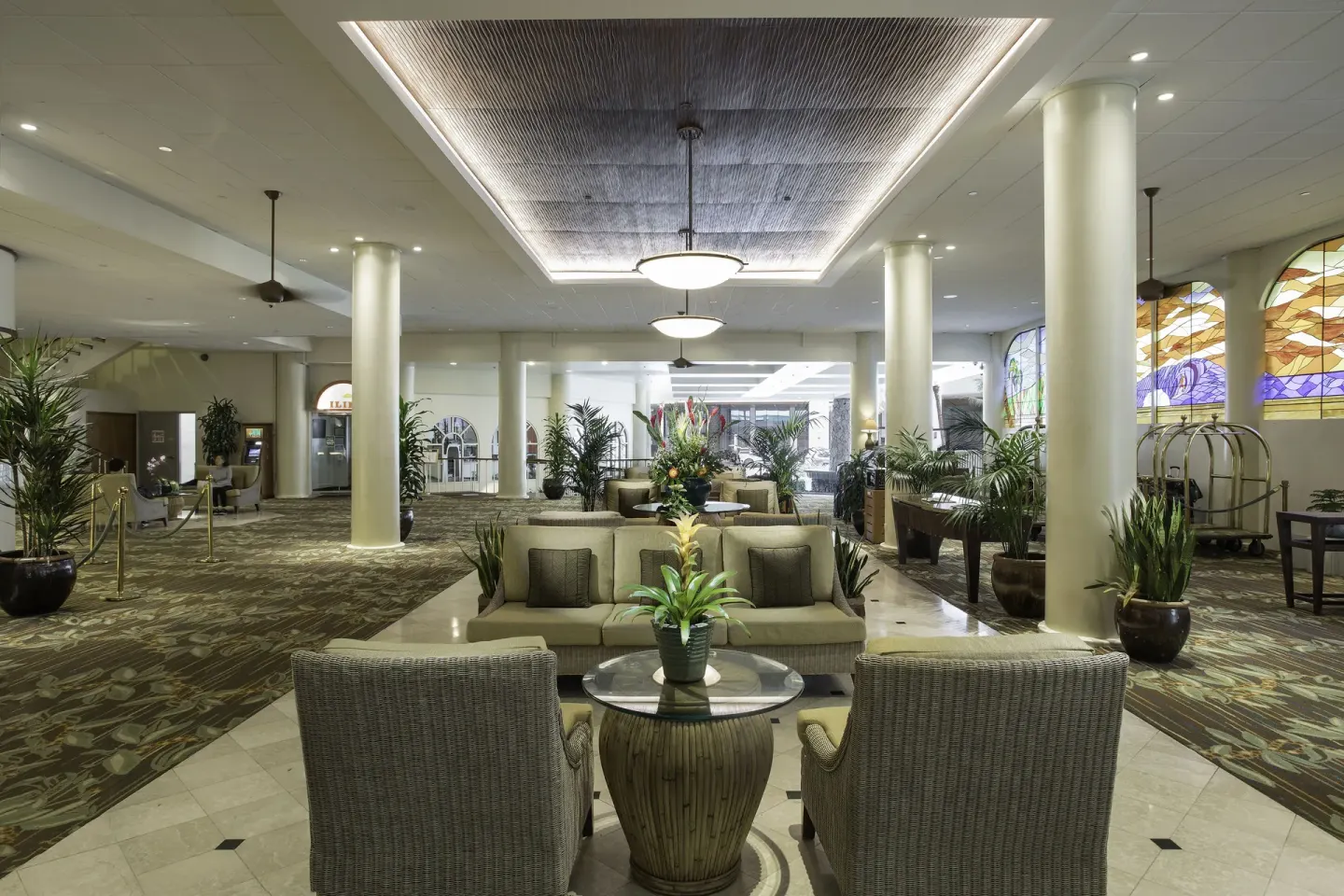 Lobby lounge area at Waikiki Resort Hotel with wicker chairs, columns, plants, and stained-glass windows.