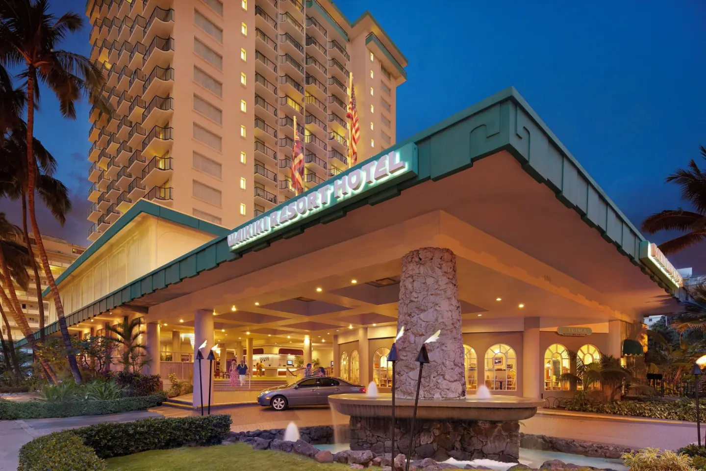 Nighttime entrance with lit hotel sign, fountain and tower at Waikiki Resort Hotel.