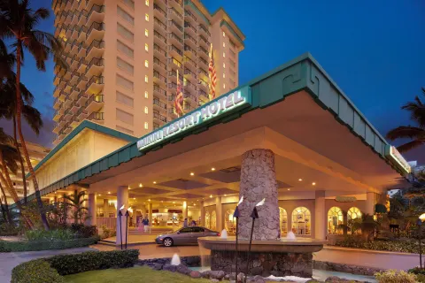 Nighttime entrance with lit hotel sign, fountain and tower at Waikiki Resort Hotel.