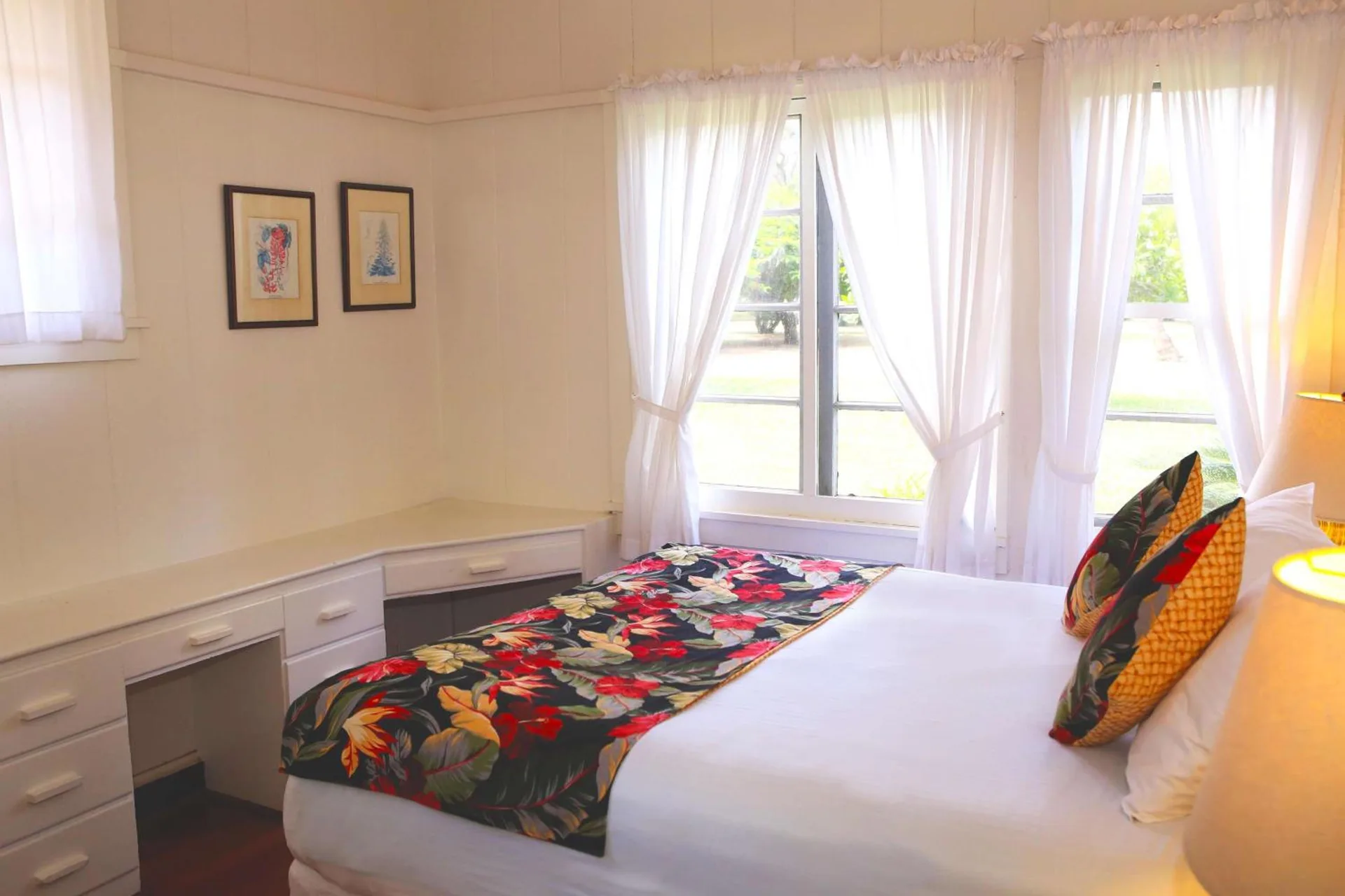 Garden-view cottage bedroom with bed, floral runner, desk, and curtained windows at Waimea Plantation Cottages.