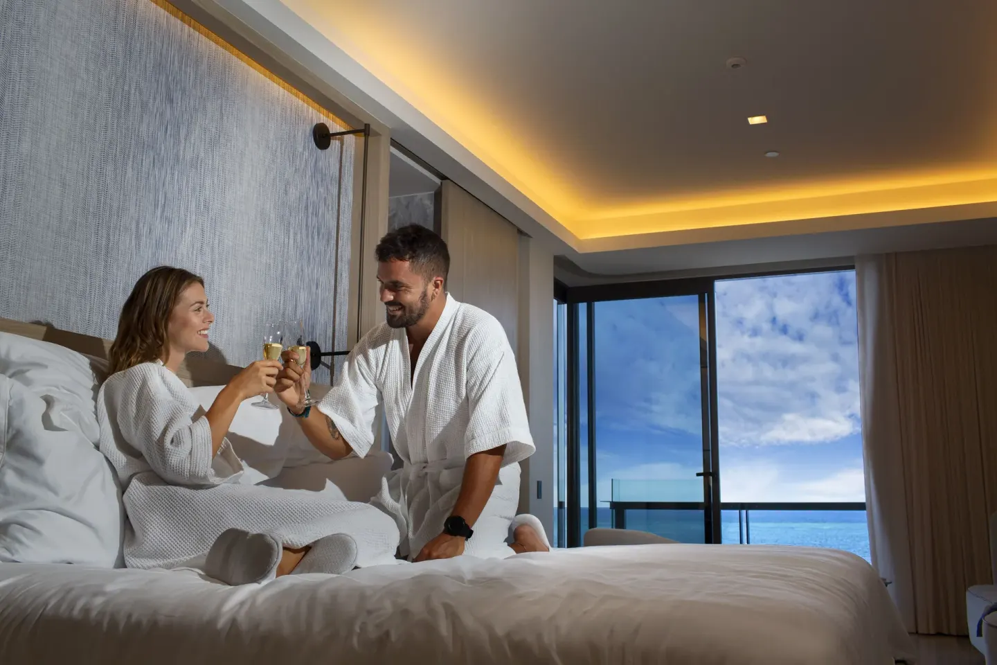 Guests toast on a king bed in an ocean-view room at Waldorf Astoria Riviera Maya.
