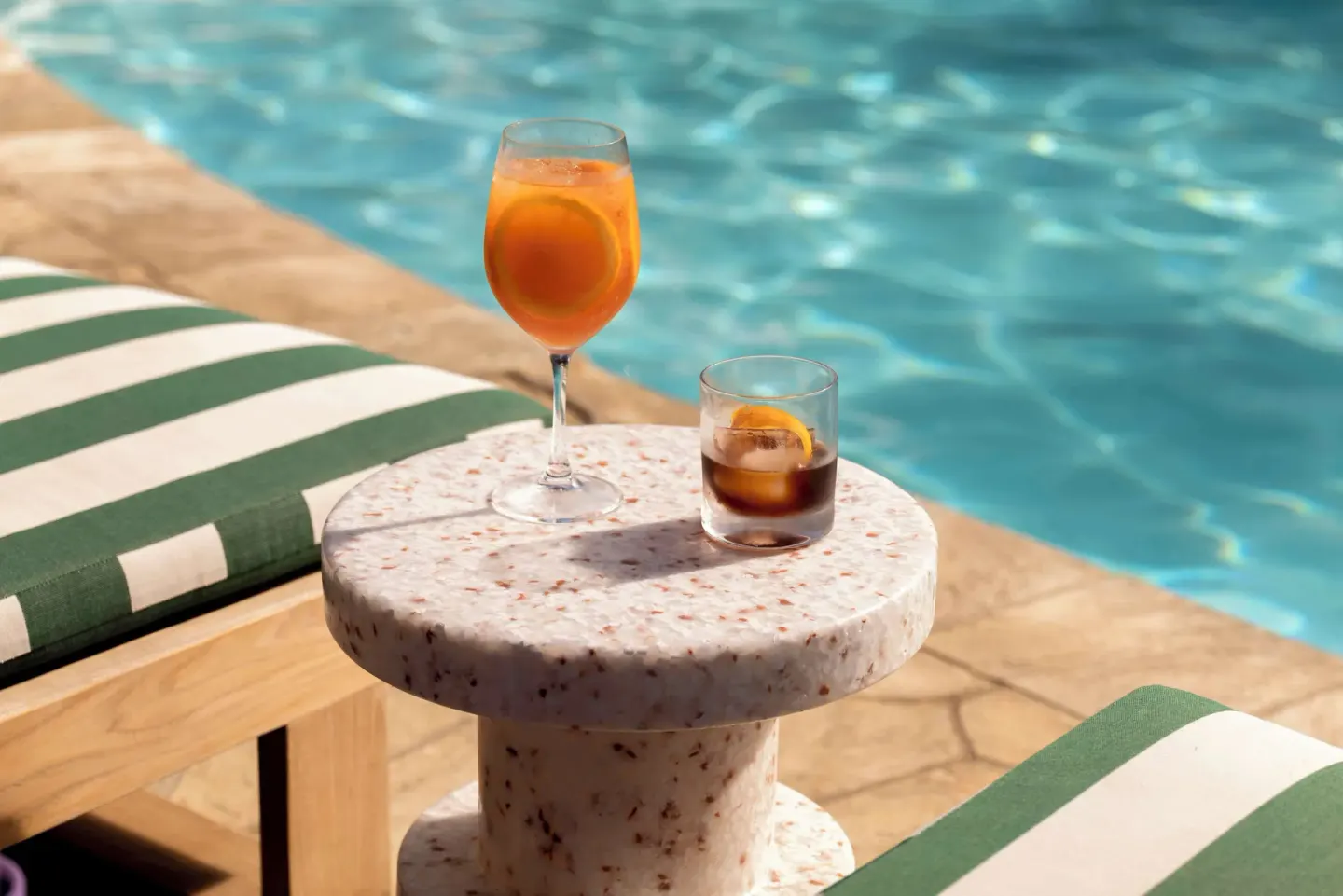 Two cocktails on a small poolside table by striped loungers at Wayfinder Waikiki.