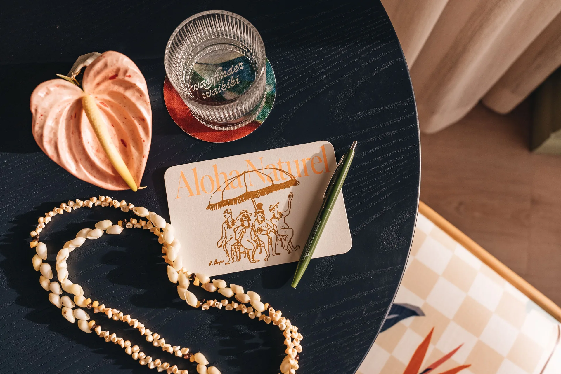 Tabletop with lei, notecard, pen, flower, and glass at Wayfinder Waikiki.